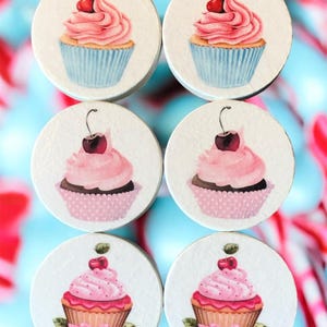 May include: Six round coasters with cupcake designs. Each coaster shows a different cupcake with pink icing and a cherry. The cupcakes are in blue, pink, and brown cases. The background is a mix of blue and red.