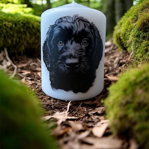 May include: A white pillar candle with a black and white dog portrait. The dog has dark fur and brown eyes. The candle is cylindrical. The background shows green moss and fallen leaves, creating a natural setting. The candle is a decorative item.