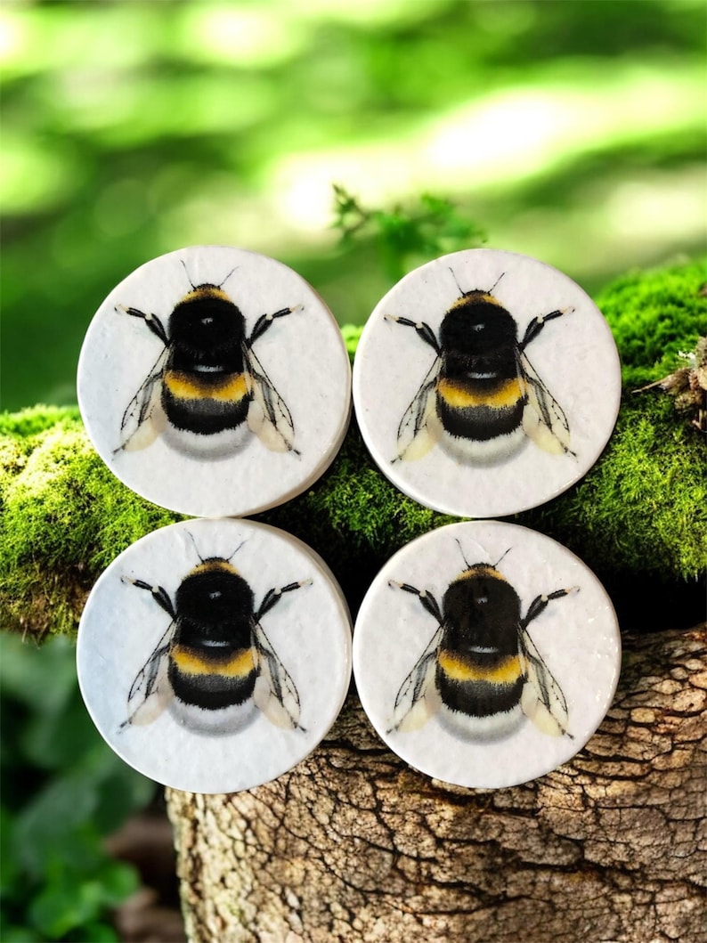 May include: Four circular coasters featuring a detailed bumblebee illustration. The bees are black, yellow, and white, set against a white background. The coasters are displayed on a textured, natural surface with a blurred green backdrop.