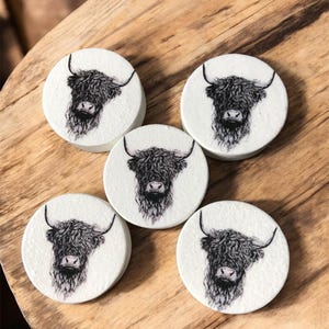 Pode incluir: Cinco bases de copos redondas brancas com uma ilustração a preto e branco da cabeça de uma vaca das Highlands. As bases de copos estão dispostas numa superfície de madeira, mostrando a arte detalhada do pelo e dos chifres do animal.