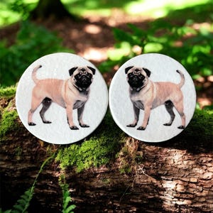 Könnte beinhalten: Zwei runde, weiße Untersetzer mit einer Illustration eines Mops. Die Möpse sind hellbraun mit schwarzen Gesichtern und Ohren. Die Untersetzer liegen auf einem moosbewachsenen Baumstamm in einer natürlichen Umgebung.