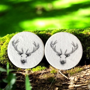 May include: Two circular white coasters featuring a black and white stag's head design. The coasters are made from a textured material and are presented on a moss-covered log, with a blurred green backdrop.