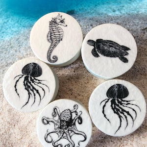 Peut inclure: Cinq dessous de verre ronds blancs avec des illustrations noires de la vie marine. Les motifs comprennent un hippocampe, une tortue, deux méduses et un poulpe. Les dessous de verre sont disposés sur une surface sablonneuse avec un arrière-plan océanique flou.