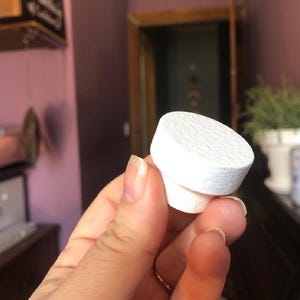 May include: A white bottle stopper with a textured top and a cylindrical base. The stopper is held in a hand, showcasing its clean design and potential use for sealing bottles. The background is a blurred interior.