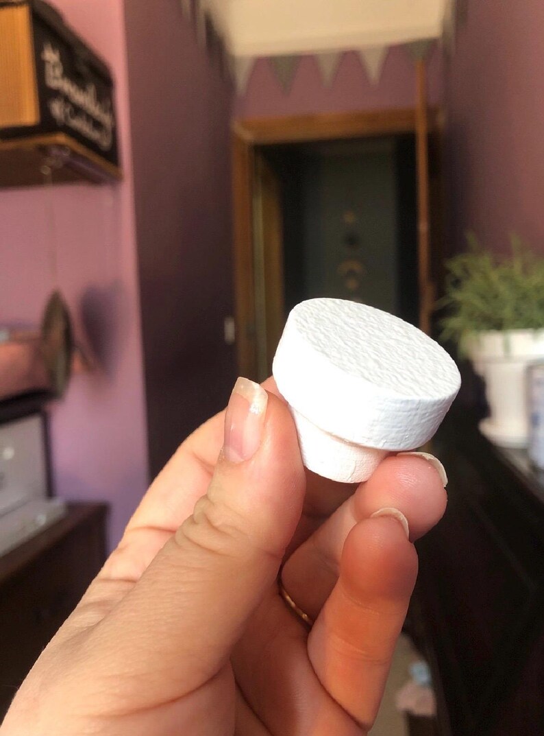 May include: A white bottle stopper with a textured top and a cylindrical base. The stopper is held in a hand, showcasing its clean design and potential use for sealing bottles. The background is a blurred interior.