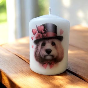 May include: White pillar candle with a dog wearing a top hat and a pink bow tie. The candle has a decorative design with hearts and roses. The candle is placed on a wooden surface.