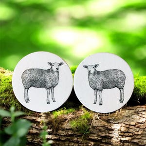 Könnte beinhalten: Zwei runde, weiße Untersetzer mit detaillierten Schwarz-Weiß-Illustrationen eines Schafs. Die Untersetzer liegen auf einem moosbewachsenen Baumstamm vor einem verschwommenen grünen Hintergrund.