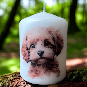 May include: A white pillar candle with a detailed illustration of a light brown and white puppy. The candle has a wick at the top and is set against a blurred green background.
