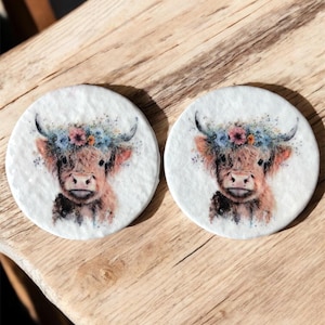Könnte beinhalten: Zwei runde, weiße Untersetzer mit jeweils einer Aquarell-Illustration einer Highland-Kuh mit Blumenkrone. Die Kühe haben braunes Fell und sind vor einem weißen Hintergrund platziert. Die Untersetzer werden auf einer Holzoberfläche präsentiert.