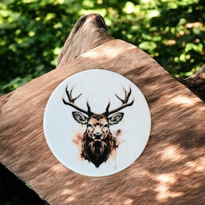 Könnte beinhalten: Ein runder, weißer Untersetzer mit einer Aquarellillustration eines Hirschkopfes und Geweihs. Der Hirsch ist in Braun- und Schwarztönen gehalten, mit einem strukturierten Aquarelleffekt. Der Untersetzer befindet sich auf einem Hintergrund aus Naturholz.