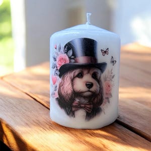 May include: White pillar candle with a dog illustration wearing a top hat and bow tie, framed by pink roses and butterflies. The candle has a single wick and is a decorative item.