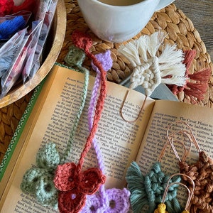 May include: A still-life image featuring a cup of coffee, open book, and handmade crochet bookmarks in various colors. The bookmarks are attached to copper paper clips. A wooden bowl with colorful embroidery floss is also present.