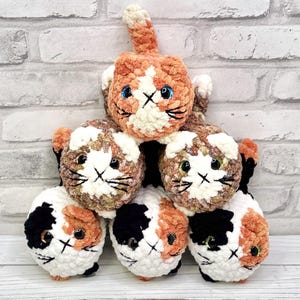 May include: A pyramid of six handmade crochet cat toys. The cats have white, orange, brown, and black fur. Each cat has a stitched face with a white muzzle, black whiskers, and blue or green eyes. The cats are arranged on a white wooden surface.