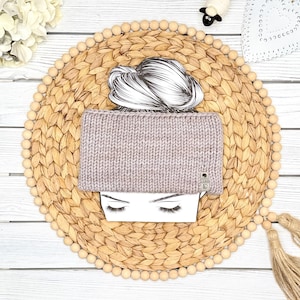 May include: A light mauve knitted headband featuring a chunky ribbed texture and a small rectangular grey brand tag on the lower right side. The headband is displayed on a white paper cutout of a head with a bun hairstyle and closed eyes.