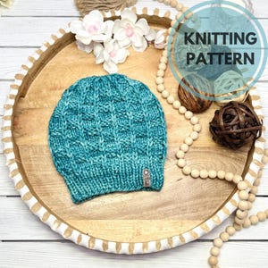 May include: A teal knitted beanie with a textured pattern sits on a wooden tray. The tray is decorated with white flowers, wooden beads, and a sign that says "KNITTING PATTERN".