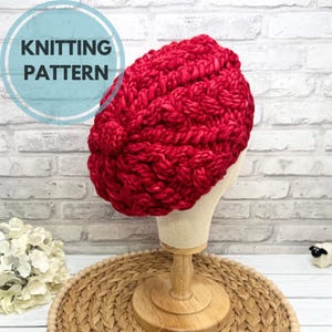 Puede incluir: Un gorro de punto rojo vibrante, exhibido en una cabeza de maniquí. El gorro presenta un diseño de punto grueso y está etiquetado como "KNITTING PATTERN" en un gráfico circular. El gorro se exhibe en un soporte de madera.
