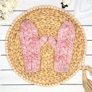 May include: A pair of chunky knit mittens in a soft dusty pink color, featuring a prominent vertical cable knit pattern throughout. The mittens have a ribbed cuff and a small rectangular tag attached to the bottom edge of the right-hand mitten.