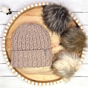 May include: A chunky knit beanie in a dusty rose color with a basketweave texture and a small rectangular tag on the cuff. Three faux fur pom-poms in shades of grey, brown, and cream are arranged nearby, along with a small black and white sheep charm.