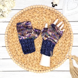 May include: A pair of fingerless gloves featuring a chunky knit texture. The lower section is a solid dark navy blue, while the upper section displays a variegated pattern of purple, brown, cream, and green tones. A small rectangular tag is attached.