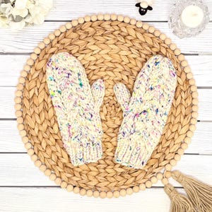 May include: A pair of thick, cream-colored knit mittens featuring a chunky cable-knit pattern and multicolored flecks of blue, pink, and yellow yarn throughout the fabric. The mittens have a ribbed cuff and a distinct thumb section on each side.