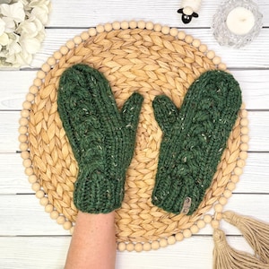 May include: A pair of forest green cable knit mittens crafted from thick, speckled yarn. The mittens feature a vertical braided cable pattern, a ribbed cuff, and a small rectangular tag attached to the lower edge of the right mitten.