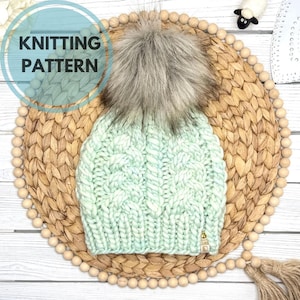 May include: A mint green cable knit beanie featuring a large, fluffy, multi-toned brown and grey faux fur pom-pom on top. The hat has a ribbed brim and a small rectangular tan tag attached to the lower right side. Text reads KNITTING PATTERN.