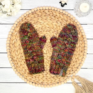 May include: A pair of chunky knit mittens featuring a variegated yarn pattern with blended shades of pink, green, orange, and purple. The mittens have a thick, textured cable knit design and a small rectangular tag attached to the lower edge.