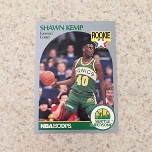 May include: A 1990 NBA Hoops rookie card featuring Shawn Kemp of the Seattle SuperSonics. The card is green, white and blue with a photo of Kemp dribbling a basketball. The card text reads "Shawn Kemp, Forward/Centre, Rookie, Seattle SuperSonics".