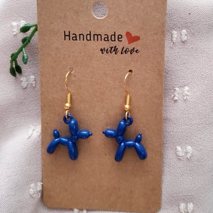 May include: A pair of blue balloon dog earrings with gold hooks. The earrings are displayed on a brown card with the text "Handmade with love".