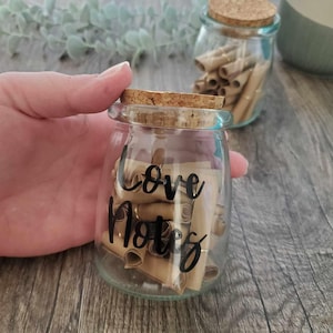 May include: A glass jar filled with rolled up pieces of paper, labeled "Love Notes". The jar has a cork lid and is sitting on a wooden surface.