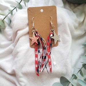 May include: A pair of silver star earrings with red, white, and blue ribbon tassels. The stars have a white outline and are decorated with small stars.