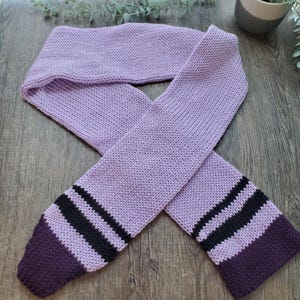 May include: A knitted scarf in a soft lavender color. The scarf features black stripes and a deep purple border at each end. The scarf is made of a soft, textured yarn, perfect for cold weather.