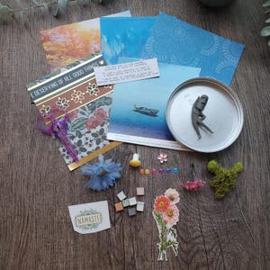 May include: A collection of craft supplies and decorative items arranged on a wooden surface. Includes patterned paper, a small figurine in a metal lid, a sticker that says "Namaste", and various embellishments like flowers and moss.