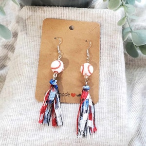 May include: Baseball-themed earrings with white and red baseball bead accents. The earrings feature silver hooks and are adorned with red, white, and blue ribbon tassels. The earrings are displayed on a brown card.