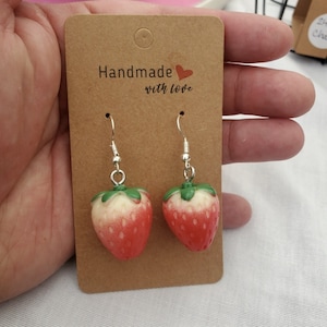 May include: A pair of silver dangle earrings with red and green strawberry charms. The earrings are on a brown card with the text "Handmade with love".