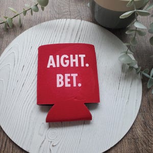 May include: A red foam beverage insulator sleeve featuring the white text AIGHT. BET. printed in a bold, sans-serif font on the front. The sleeve is designed to fit around a standard can or bottle to provide insulation and a comfortable grip.