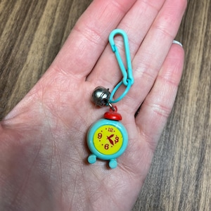 May include: A small, turquoise alarm clock charm with a red top and yellow clock face. The clock shows the time as 6:00. The charm is attached to a turquoise clasp and a small silver bell.