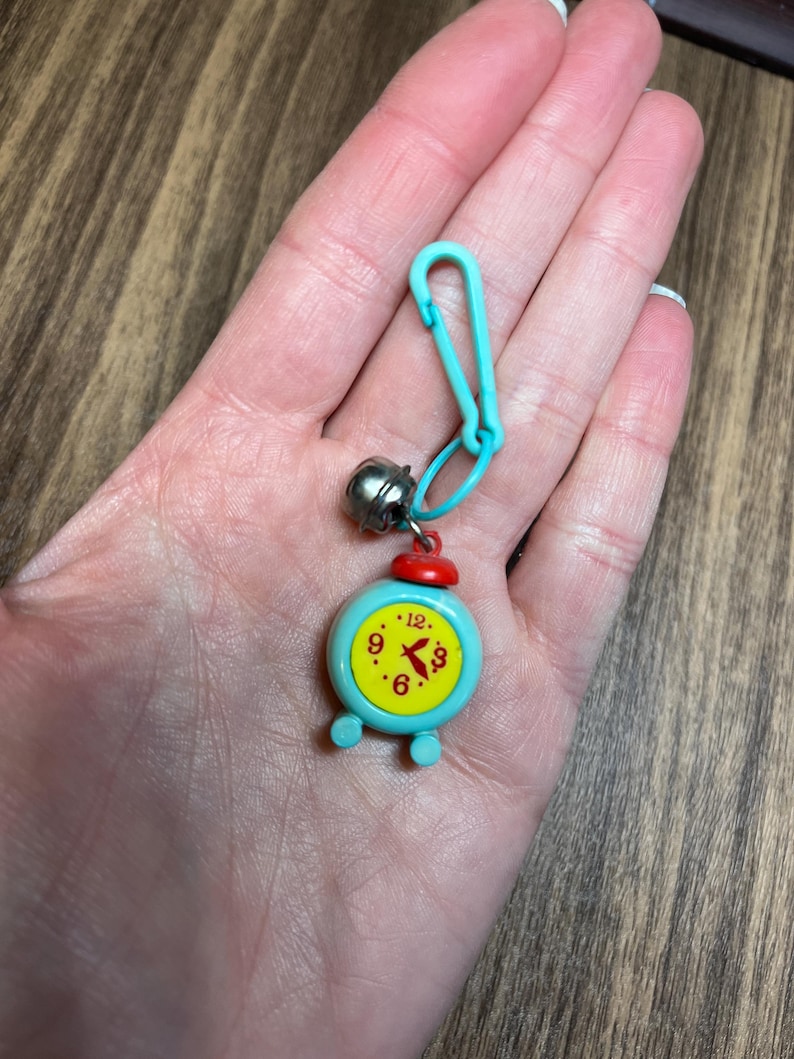 May include: A small, turquoise alarm clock charm with a red top and yellow clock face. The clock shows the time as 6:00. The charm is attached to a turquoise clasp and a small silver bell.