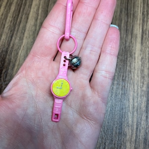 May include: A pink toy watch with a yellow face and black hands, attached to a pink clip and a silver bell. The watch has a rectangular shape and is held in a hand.