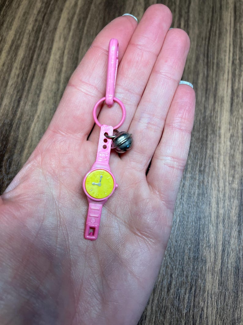 May include: A pink toy watch with a yellow face and black hands, attached to a pink clip and a silver bell. The watch has a rectangular shape and is held in a hand.