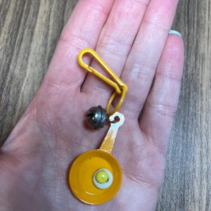 May include: A yellow keychain with a miniature frying pan charm featuring a fried egg. The charm is connected to a yellow clip and a small silver bell. The frying pan is yellow with a white and yellow egg.