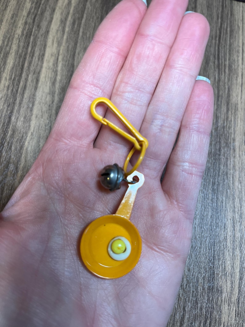 May include: A yellow keychain with a miniature frying pan charm featuring a fried egg. The charm is connected to a yellow clip and a small silver bell. The frying pan is yellow with a white and yellow egg.