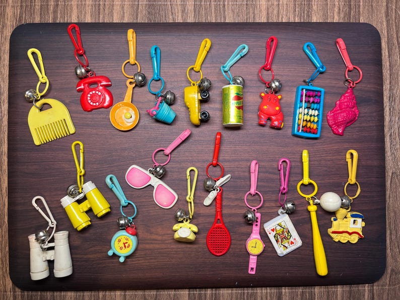 May include: A collection of colorful keychains with various miniature designs, including a comb, phone, frying pan, sunglasses, and a tennis racket. Each keychain has a metal clasp and a small bell. The keychains are arranged on a dark wooden surface.