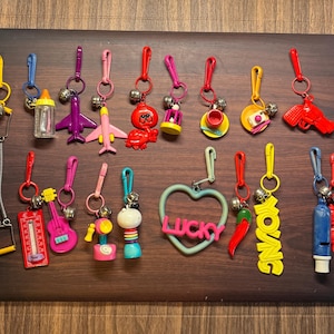 May include: A collection of colorful keychains with various designs, including a baby bottle, airplanes, a red gun, a guitar, and a heart-shaped charm with the word "LUCKY". Each keychain has a metal clip and a small bell.