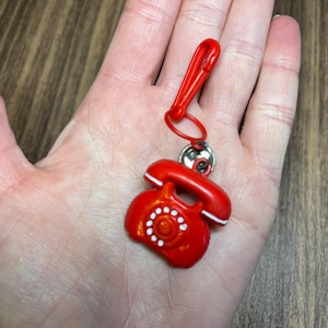 May include: A red telephone charm with a matching red clip. The vintage-style phone has white accents and a circular dial. The charm is attached to a silver-colored ring.