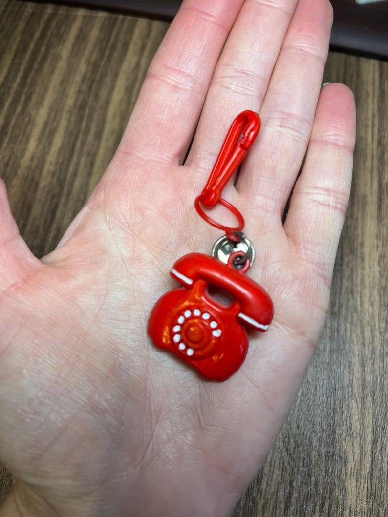 May include: A red telephone charm with a matching red clip. The vintage-style phone has white accents and a circular dial. The charm is attached to a silver-colored ring.