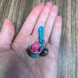 May include: A miniature charm featuring a pink creature in a blue bucket, attached to a blue clip with a small silver bell. The charm is held in a hand against a wooden background.