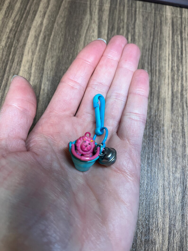 May include: A miniature charm featuring a pink creature in a blue bucket, attached to a blue clip with a small silver bell. The charm is held in a hand against a wooden background.