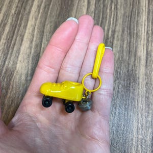 May include: A yellow roller skate keychain with black wheels and a silver bell charm. The keychain is held in a hand against a wood grain background. The roller skate is a miniature replica.