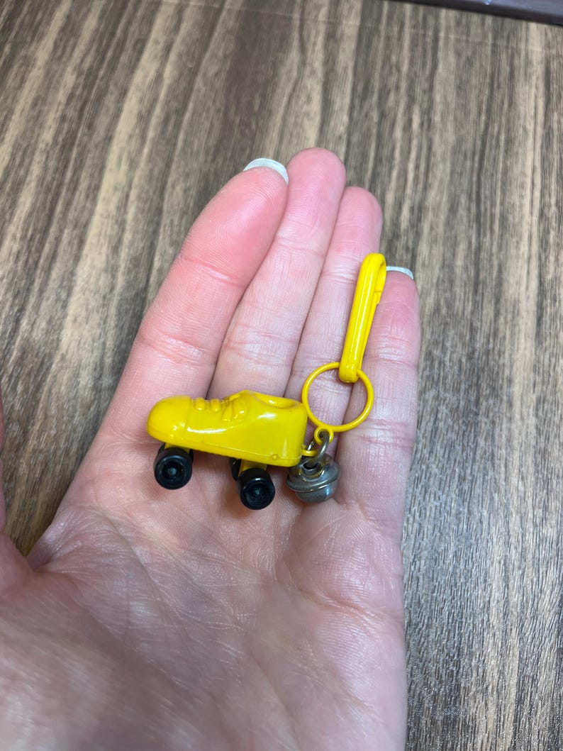 May include: A yellow roller skate keychain with black wheels and a silver bell charm. The keychain is held in a hand against a wood grain background. The roller skate is a miniature replica.